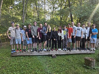 Gruppenfoto mit Jugendlichen, die eng zusammen auf einem Holzsteg im Wald stehen.