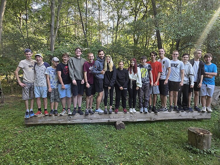 Gruppenfoto mit Jugendlichen, die eng zusammen auf einem Holzsteg im Wald stehen.