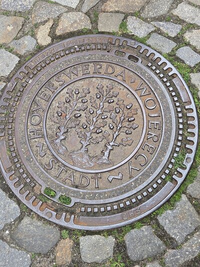 Ein Kanaldeckel zwischen Pflastersteinen. Auf dem Deckel steht "Stadt Hoyerswerda Wojerecy" und in der Mitte sind drei Eichenbäume abgebildet.