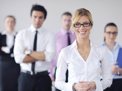 Eine Frau in einem weißen Hemd und mit Brille steht im Vordergrund und lächelt. Hinter ihr sind mehrere Personen, die verschwommen sind und Geschäftskleidung tragen, was auf ein professionelles Umfeld hinweist.