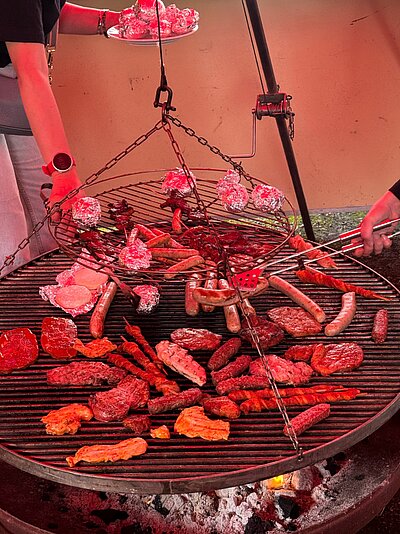 Auf einem Schwenkgrill liegen verschiedene Fleischspezialitäten wie Grillsteaks, Würstchen, Grillfackeln, Putensteaks, aber auch Grillkäse und Kartoffeln in Alufolie. Eine Person ist gerade dabei, die Folienkartoffeln auf den Grill zu legen, während eine andere die Würstchen wendet.