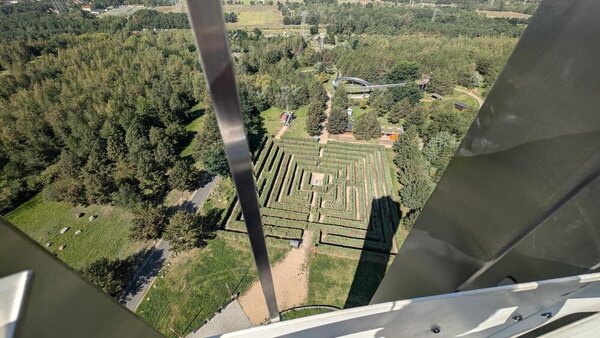 Der Blick zeigt von einem hohen Turm durch ein Metallgitter auf den darunter liegenden Irrgarten sowie eine Sommerrodelbahn. Außen herum sind Wiesen und Wälder.