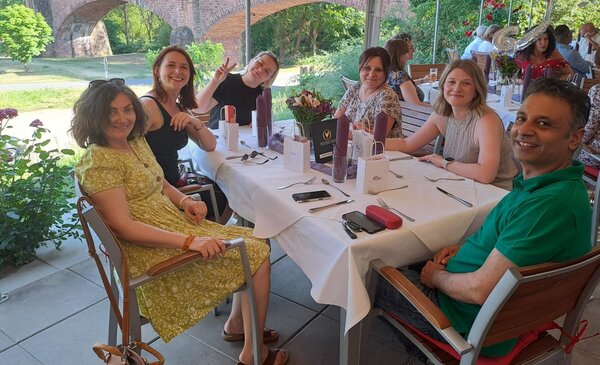 Eine Gruppe von sechs Personen sitzt an einem langen, festlich gedeckten Tisch auf deiner Terrasse mit Blick auf einen grünen Garten und historische Backsteinbögen. Auf dem Tisch stehen Blumen, Gläser und Geschenktüten. Alle Personen lächeln und eine macht das Peacezeichen.
