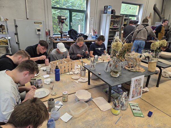 Eine Gruppe junger Menschen sitzt um einen großen Holztisch in einer Werkstatt. Die jungen Menschen bemalen Teller und Tassen. Auf dem Tisch stehen weitere Bastelmaterialien und Blumensträuße. Im Hintergrund sind weitere junge Menschen an Werkbänken zu sehen.