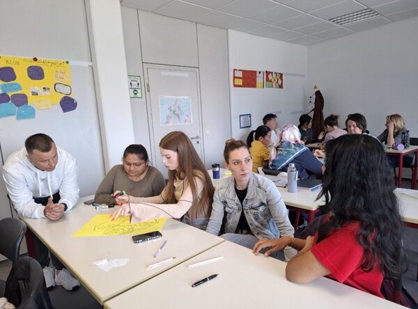 Weibliche und männliche Teilnehmende des B2-Kurses sitzen im Unterricht an Tischen und arbeiten gemeinsam an ihren Präsentationen.