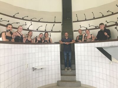 Eine Gruppe von neun Personen steht hinter einem podestartigen Rondell mit weiß gekachelten Wänden. An jedem Platz und auch auf den Plätzen in den höheren Etagen dahinter sind schwarze Halterungen oder Lampen angebracht. Die Personen posieren lächelnd oder mit Peace-Zeichen für die Kamera. Ein Mann steht in der Mitte der Treppe und blickt direkt in die Kamera.