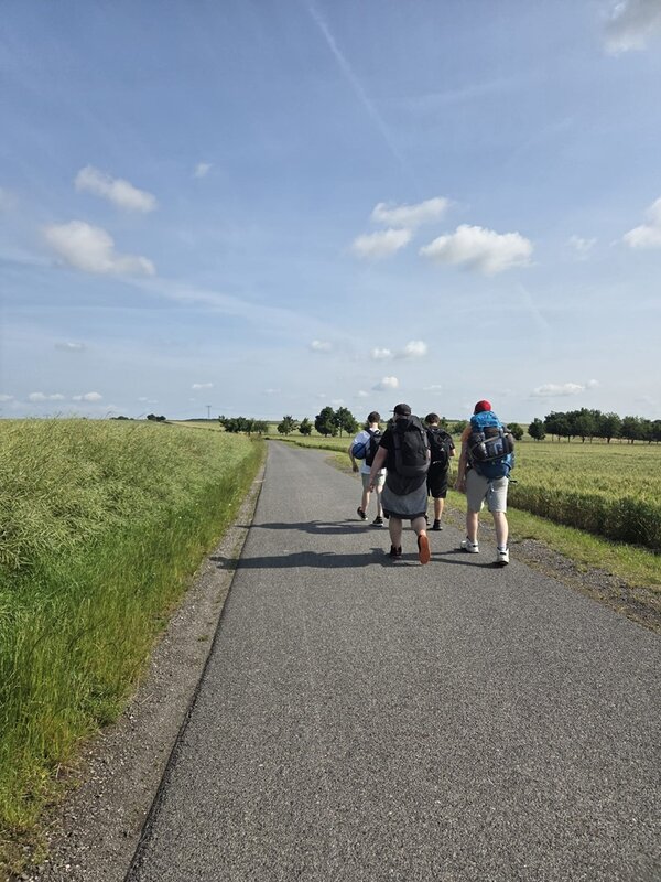 Die Auszubildenden zu Fachlageristen wandern mit ihren Rucksäcken eine Straße entlang, das Wetter ist sommerlich, der Himmel blau, links und rechts weite Felder.