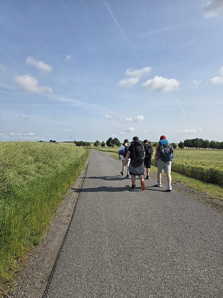 Pilgern mit Perspektive Die Auszubildenden zu Fachlageristen wandern mit ihren Rucksäcken eine Straße entlang, das Wetter ist sommerlich, der Himmel blau, links und rechts weite Felder.