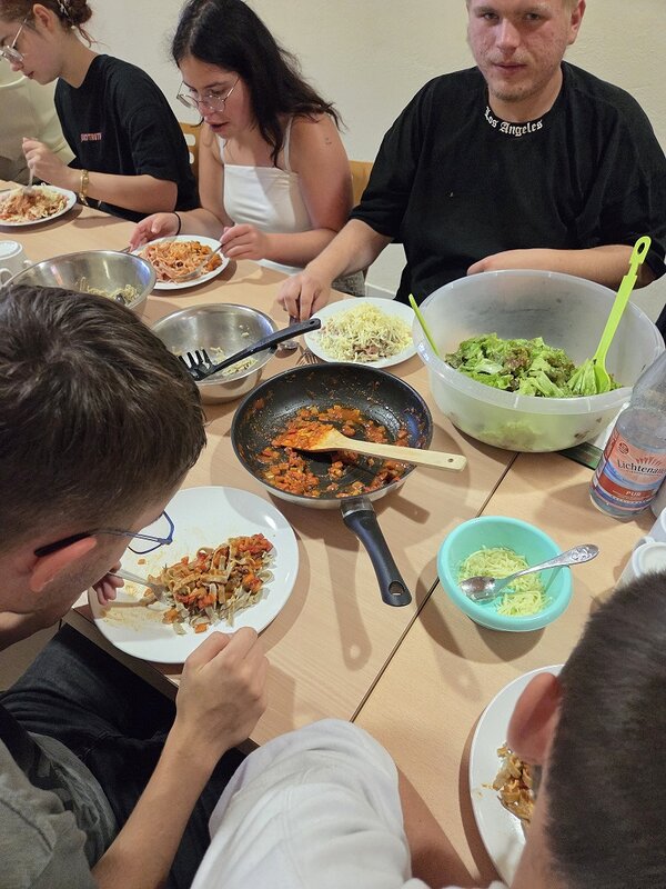 Eine Gruppe von Menschen sitzt an einem Tisch und isst gemeinsam. Auf dem Tisch stehen Schüsseln mit Nudeln, Salat und anderen Speisen.