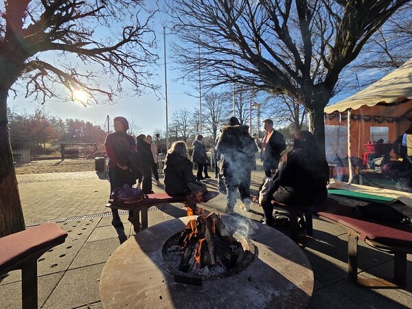 Eine Gruppe Menschen sitzt um eine Feuerstelle im Freien, im Hintergrund ein Zelt, Bäume und ein strahlend blauer Himmel
