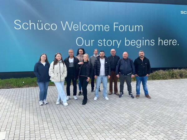 Gruppenbild draußen vor einer blauen Wand, auf der steht "Schüco Welcome Forum - Our story begings here."