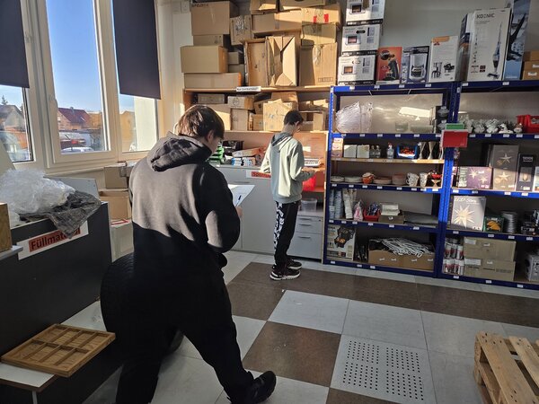 Zwei Jugendliche arbeiten in einem hellen Lager- und Verkaufsraum zwischen Regalen mit Waren und Kartons; eine Person hält Unterlagen in der Hand, die andere steht am Regal.