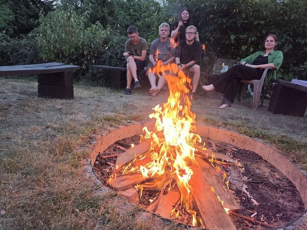 Eine Gruppe von fünf Personen sitzt entspannt auf Holzbänken um ein Lagerfeuer in einem grünen Garten. Das Feuer brennt lebhaft in einer feuerfesten Grube, während die Personen sich unterhalten und die Atmosphäre genießen.