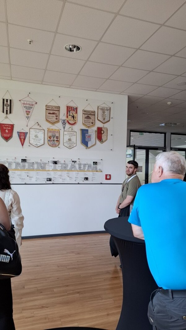 Ein Guide erklärt Exponate im Inneren des Vereinsgebäudes; im Hintergrund hängen historische Vereinswimpel an der Wand, während die Besucher aufmerksam zuhören.