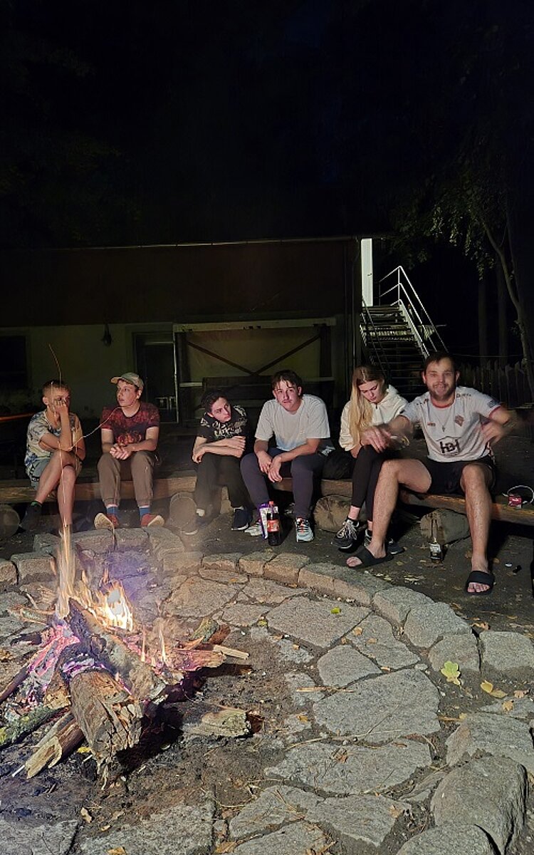 Gruppe Jugendlicher sitzt abends um ein Lagerfeuer, im Vordergrund brennende Flammen.