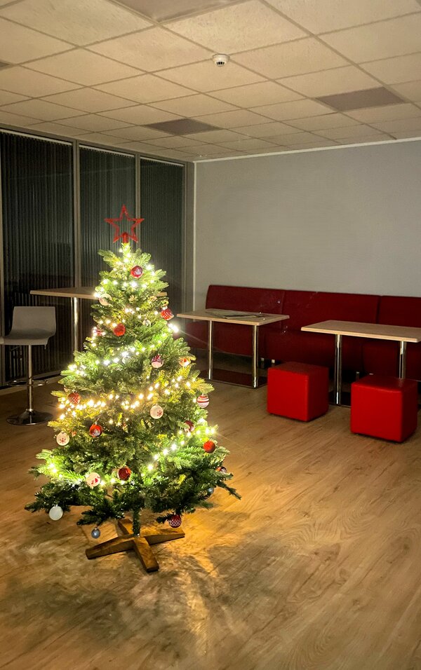 Kleiner Aufenthaltsbereich mit leuchtendem Weihnachtsbaum im Vordergrund, dahinter rote Sitzbänke, Tische und rote Würfelhocker.