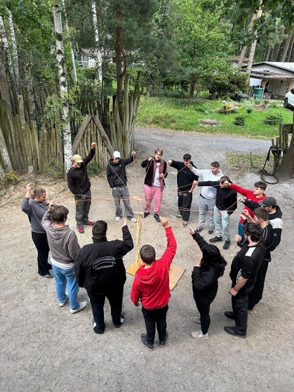 Auf einem Platz im Freien steht eine Gruppe junger Menschen im Kreis. Jede Person hält einen Faden in der Hand und die Fäden laufen in der Mitte zusammen.