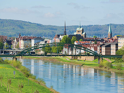 Minden in Nordrhein-Westfalen Im Vordergrund die Weser mit grünem Ufer, im Hintergrund ein Teil von Minden vor einem bewaldeten Hügel.