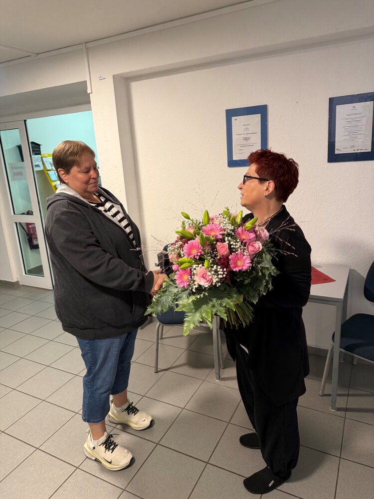 Zwei Frauen stehen sich in den Räumen der Euro-Schulen gegenüber und schütteln sich die Hand. Eine der Frauen hält einen großen Blumenstrauß mit rosa Rosen und Gerbera.