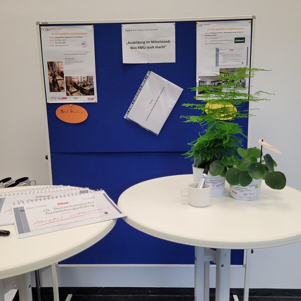 Blaues Flipchart im Hintergrund mit Informationen zum RAD, davor zwei Stehtische, auf einem davon stehen zwei Grünpflanzen.