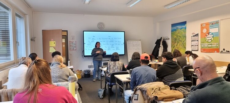 Unterrichtsszene in einem hellen Klassenraum: Eine Lehrkraft steht vor einer digitalen Tafel mit handschriftlichen Notizen und erklärt den Lernstoff, während die erwachsenen Teilnehmenden aufmerksam an ihren Tischen sitzen und mitschreiben. Der Raum ist mit Whiteboard, Flipchart, Wandkarten und Lehrmaterialien ausgestattet. Die Lernenden arbeiten konzentriert in einer ruhigen Kursatmosphäre. Das Bild steht für Erwachsenenbildung, Sprachkurs oder Weiterbildung im Klassenverband und vermittelt praxisnahen Präsenzunterricht in einem modernen Raum an den Euro-Schulen Potsdam.