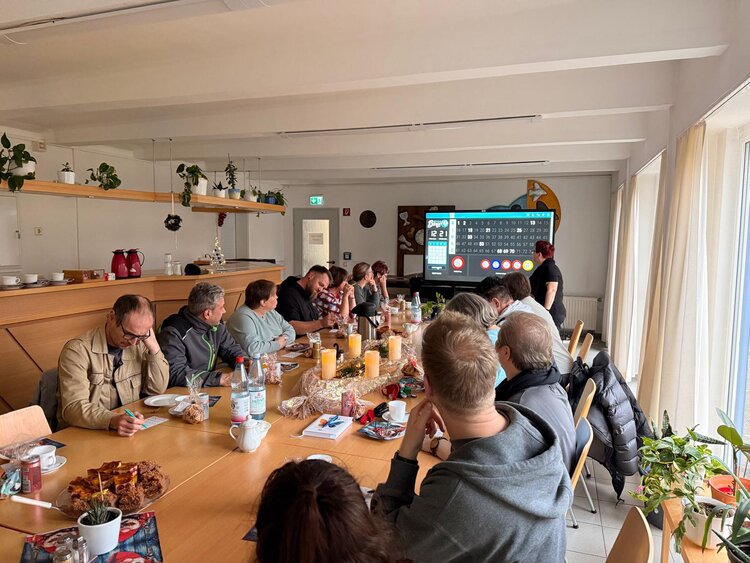 Eine Gruppe von Menschen sitzt an einem langen Tisch in einem hellen Raum und spielt gemeinsam Bingo. Auf dem Tisch stehen Tassen, Getränke und Gebäck, im Hintergrund ist eine digitale Bingo-Anzeige zu sehen.