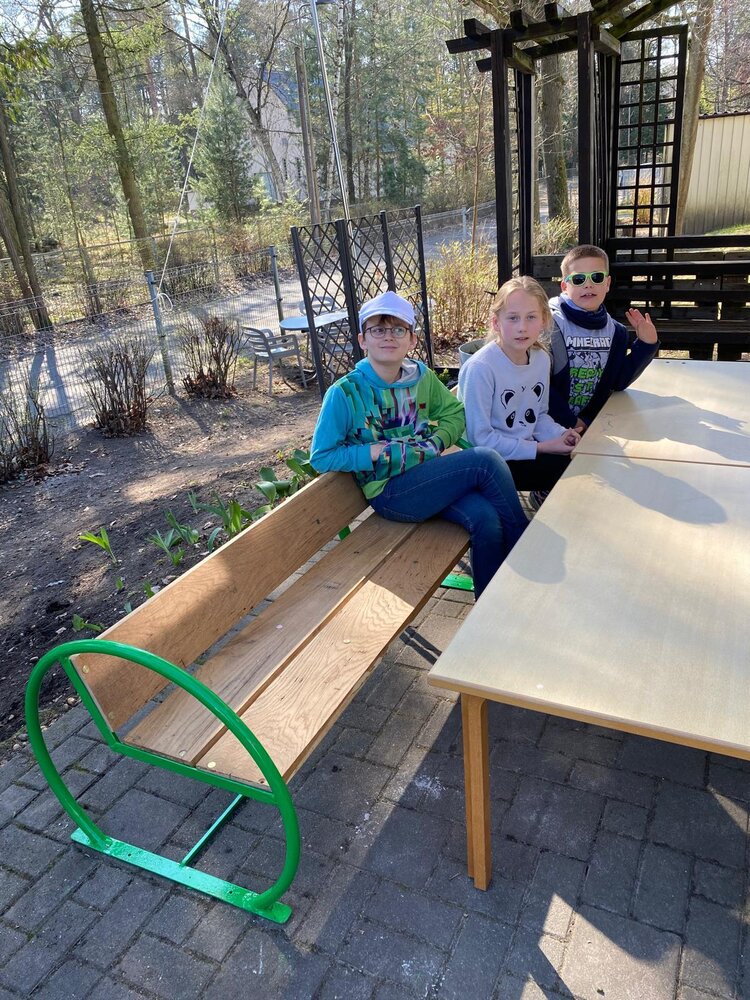 Drei Kinder sitzen auf einer Holzbank an einem Tisch im Gartenbereich und schauen in die Kamera.