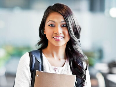 Eine junge Frau mit langen, dunklen Haaren und einem natürlichen Lächeln steht in einem hellen Innenraum. Sie trägt einen Rucksack über den Schultern und hält eine braune Mappe oder ein Heft vor sich. Ihr Oberteil ist hell und locker geschnitten. Der Hintergrund ist unscharf, aber es scheint sich um einen öffentlichen Raum wie eine Schule oder ein Büro zu handeln.