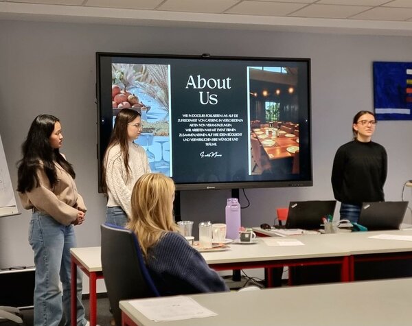 Zwei Personen stehen vorne im Raum und präsentieren vor einer Gruppe von Zuhörenden. Auf dem Bildschirm hinter ihnen ist eine Webseite mit der Überschrift "About Us" und einem Text auf Deutsch zu sehen, der die Philosophie eines Unternehmens beschreibt, das sich auf Catering in verschiedenen Veranstaltungsarten spezialisiert hat. Eine Person sitzt im Vordergrund und hört der Präsentation zu. Der Raum ist ein Seminar- oder Klassenraum mit Tischen und Stühlen.