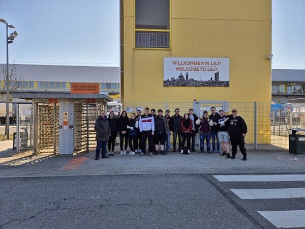 Eine Gruppe von jungen Menschen stehen auf der gegenüberliegenden Seite einer Straße und lächeln in die Kamera. Auf der rechten Seite befindet sich ein Zebrastreifen.