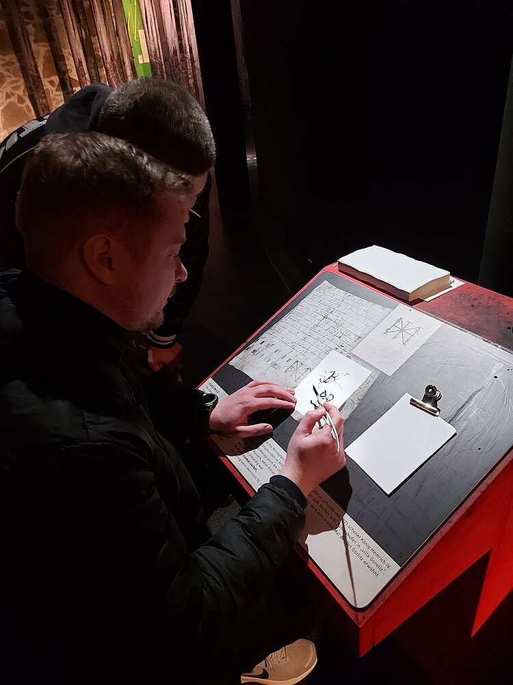 Ein männlicher Teilnehmer sitzt an einem alten Tisch im Museum und schreibt, wie in alten Zeiten, mit einer Feder auf ein Blatt Papier.