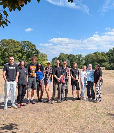 Eine Gruppe von elf jungen Erwachsenen steht bei sonnigem Wetter auf einer trockenen Wiese in einem Park. Im Hintergrund sind zahlreiche Bäume zu sehen, einige Personen halten Badmintonschläger in der Hand.