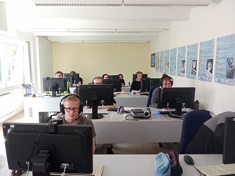 Bericht zur Berufsschulschließung am 08.05.2025 Ein heller, moderner Schulungsraum mit mehreren Reihen an Tischen, ausgestattet mit Computern und Monitoren. Junge Erwachsene sitzen mit Kopfhörern vor den Bildschirmen und konzentrieren sich auf ihre Aufgaben. An der Wand hängen informative Plakate.