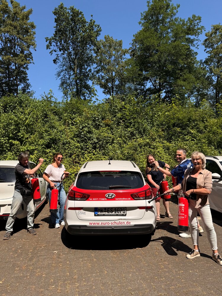 Fünf lachende Personen mit Feuerlöschern stehen um ein rot-weißes Firmenauto der Euro-Schulen Iserlohn. Das Auto steht auf einem Parkplatz mit Büschen und Bäumen im Hintergrund.