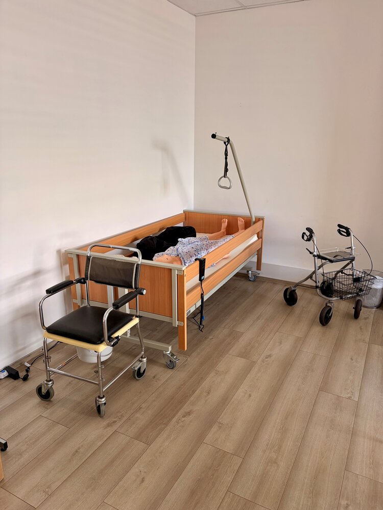Praxisraum mit Pflegebett, Übungspuppe, Toilettenstuhl und Rollator für die praktische Ausbildung im Pflege- und Gesundheitsbereich.