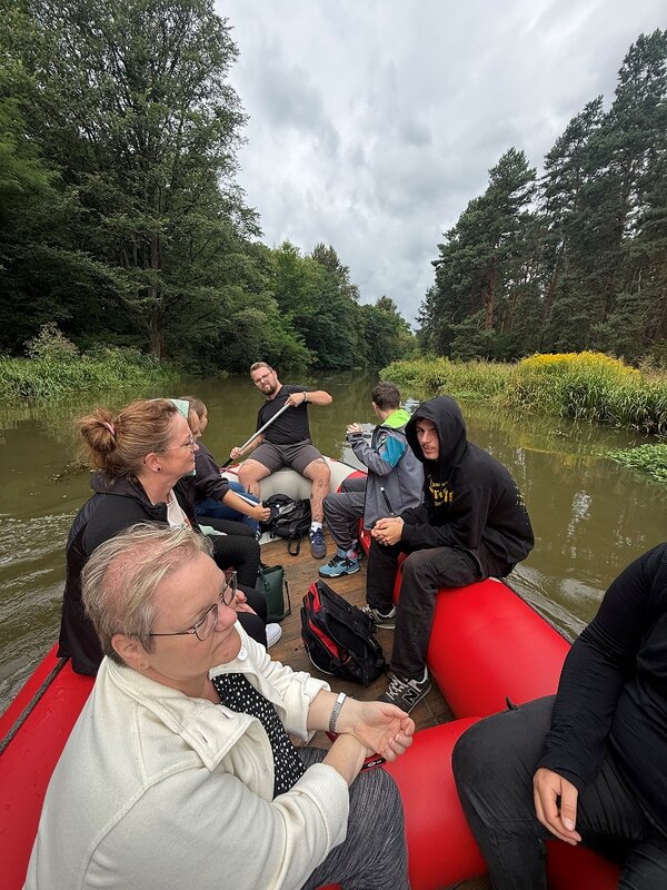 Eine Gruppe von Menschen sitzt in einem roten Schlauchboot auf einem ruhigen Fluss, umgeben von üppigem Grün und Bäumen. Die Stimmung ist entspannt, und einige Personen unterhalten sich oder schauen sich die Umgebung an, während eine Person paddelt.