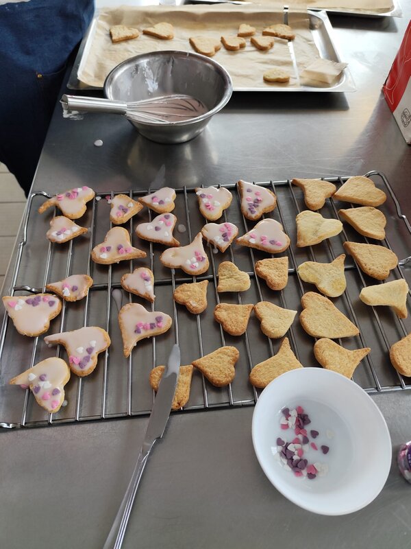 Herzförmige Kekse werden auf einem Gitterrost glasiert und mit kleinen bunten Zuckerherzen dekoriert. Eine Schüssel mit Dekorationselementen und ein Schneebesen liegen daneben.