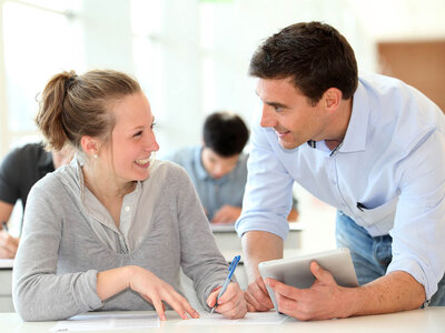 Eine Frau mit einem grauen Pullover und einem Mann mit einem hellblauen Hemd sitzen an einem Tisch. Die Frau schreibt auf ein Blatt Papier, während der Mann ein Tablet hält. Beide lächeln und wirken engagiert in ihrer Arbeit. Im Hintergrund sind weitere Personen zu sehen, die ebenfalls an Tischen arbeiten.