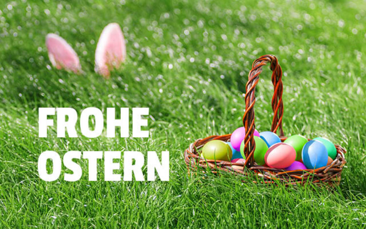 Auf einer Wiese steht ein Korb mit bunten Eiern darin, etwas weiter hinten sind die Ohren eines Hasen erkennbar, die aus dem Gras herausragen. Auf dem Bild steht in weißen Lettern "Frohe Ostern".