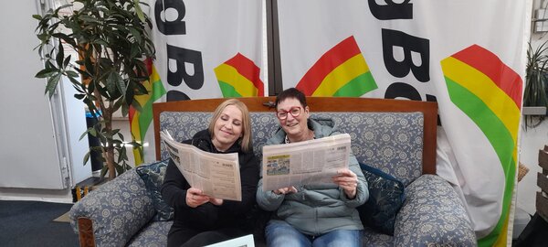 Zwei Frauen sitzen gemütlich auf einem Sofa, lesen Zeitungen und lächeln, während im Hintergrund große Stoffbanner in Rot, Gelb und Grün hängen.