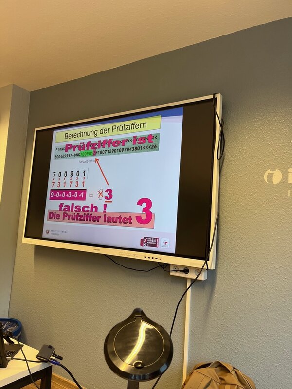 Ein großer Monitor zeigt während einer polizeilichen Schulung zur Dokumentenfälschung die Berechnung einer Prüfziffer in Ausweisdokumenten. Zu sehen sind Beispielzahlen, farbige Markierungen und ein Rechenschritt zur Kontrolle der Richtigkeit. Vor dem Bildschirm stehen technische Geräte, darunter eine Dokumentenkamera. Die Schulung findet in einem Klassenraum der Euro-Schulen Hannover statt.