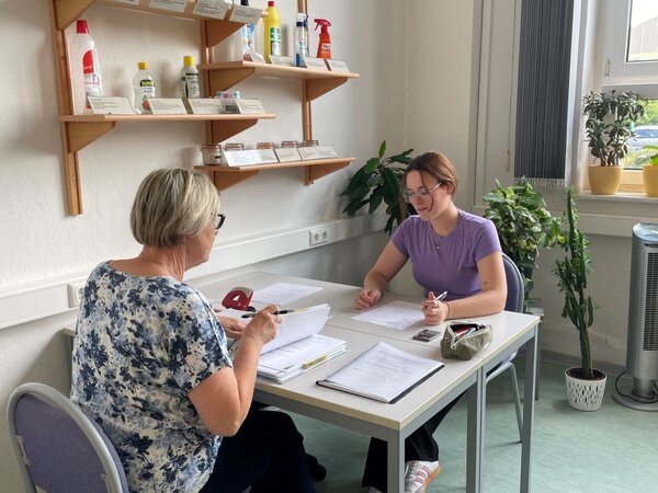 Zwei Personen sitzen an einem hellen Tisch in einem Büro oder Beratungsraum. Die ältere Person mit kurz geschnittenen, hellen Haaren und die jüngere Person in lila T-Shirt unterhalten sich und bearbeiten gemeinsam Dokumente. Im Hintergrund sind Regale mit Produkten zum Putzen und Pflanzen zu sehen.
