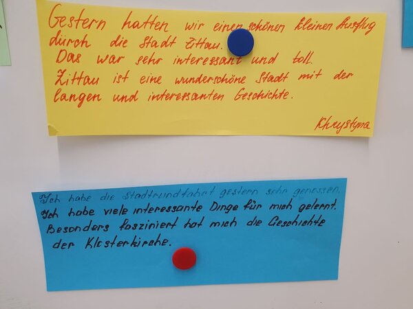 Zwei handgeschriebene Feedback-Karten sind an einer Pinnwand befestigt. Die obere Karte auf gelbem Papier beschreibt eine positive Erfahrung in Zittau, während die untere Karte auf blauem Papier eine weitere positive Rückmeldung über eine Stadtführung gibt.