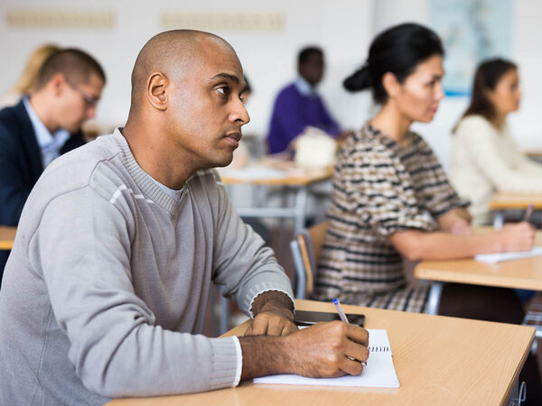 Im Vordergrund sitzt ein Mann mit Block und Stift an einem Tisch und schaut aufmerksam nach vorne; im Hintergrund sind weitere Menschen an Schultischen zu sehen.