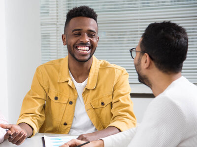 Zwei Männer sitzen an einem Tisch in einem Büroumfeld. Der Mann links, mit kurzem Afro-Haarschnitt und einem gelben Jackett, lächelt breit und scheint sich in einem angeregten Gespräch zu befinden. Der Mann rechts, mit Brille und weißem Pullover, hält einen Stift und scheint Notizen zu machen oder auf ein Dokument zu zeigen. Beide wirken freundlich und engagiert.