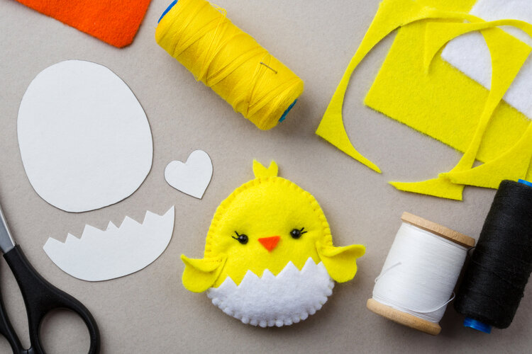 Symbolbild eines Küken, das aus gelbem und weißen Filzstoff genäht wurde. Auf dem Tisch liegen außerdem eine Papiervorlage, eine Schere und Rollen mit unterschiedlichen Fäden.