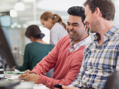 Zwei Männer sitzen lächelnd nebeneinander in einem Büro und arbeiten gemeinsam an einem Computer. Der Mann links trägt einen roten Pullover über einem weißen Hemd und wirkt fröhlich, während er auf den Bildschirm schaut. Der Mann rechts, in einem karierten Hemd, lacht ebenfalls und scheint sich über etwas zu amüsieren. Im Hintergrund sind weitere Kollegen zu sehen, die ebenfalls an ihren Arbeitsplätzen sitzen. Die Szene wirkt lebendig und kollegial, was auf eine positive Arbeitsatmosphäre hindeutet.
