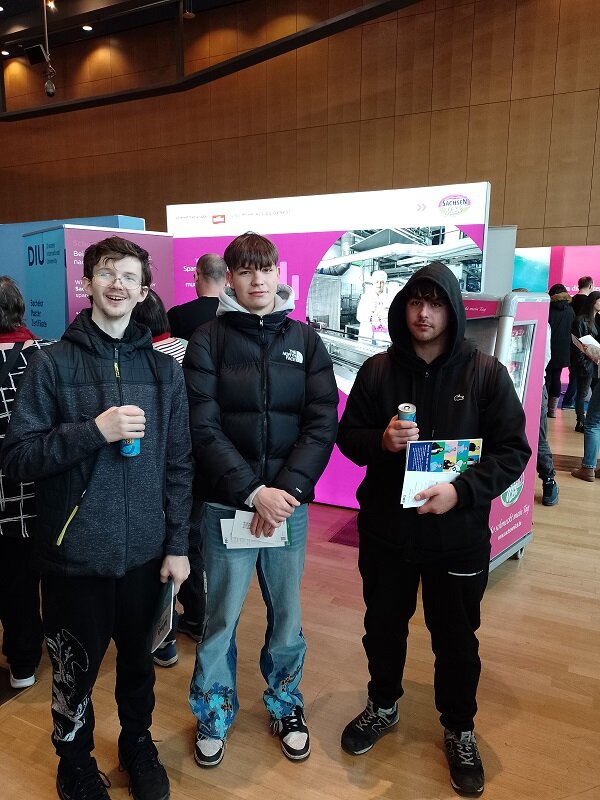 Drei junge Messebesucher mit Getränken und Infoflyern lächeln in die Kamera; hinter ihnen ein farbiger Messestand, viele Menschen.