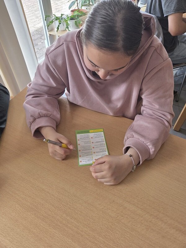 Eine Person in einem rosafarbenen Hoodie sitzt an einem Tisch und füllt einen Fragebogen oder ein Arbeitsblatt mit einem Stift aus. Der Fokus liegt auf den Händen und dem Dokument.
