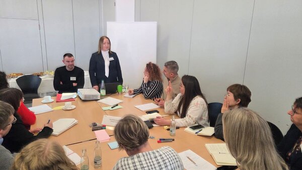 Eine Frau mit Namensschild in einem Seminarraum steht neben einem sitzenden Mann am Ende eines Tisches und spricht zu einer Gruppe von Personen, die um den Tisch herum sitzen. Die Teilnehmenden wirken interessiert und machen sich Notizen. Auf dem Tisch liegen Unterlagen, Stifte und Getränke. Im Hintergrund ist eine Leinwand zu sehen.